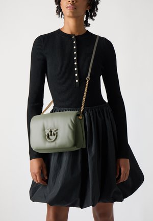Grüne Leder-Crossbody-Tasche mit strukturiertem Finish, ausgestattet mit einem dekorativen Logo und einem goldenen Kettenriemen. Der Hintergrund ist neutral.