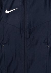 Granatowa kurtka z kapturem z zamkiem z przodu, z białym logo Nike na lewej piersi. Materiał wydaje się lekki i ma teksturowane wykończenie.