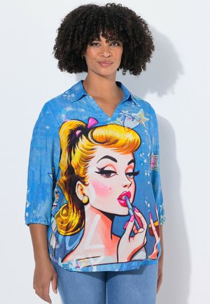 Kobieta z kręconymi włosami, ubrana w niebieską koszulę, na której znajduje się duża, kolorowa ilustracja w stylu pop-art przedstawiająca blondynkę nakładającą pomadkę.