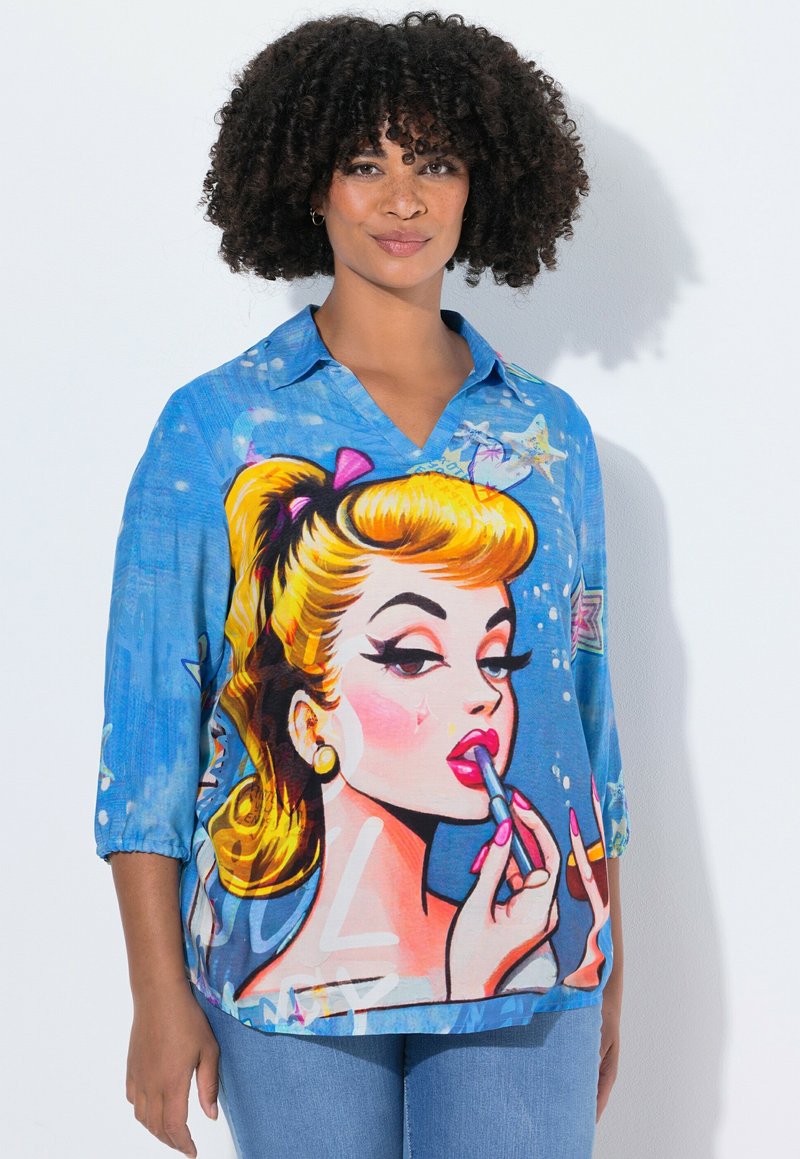Femme avec des cheveux bouclés portant une chemise bleue ornée d'une grande illustration colorée au style pop art d'une femme blonde en train de s'appliquer du rouge à lèvres.