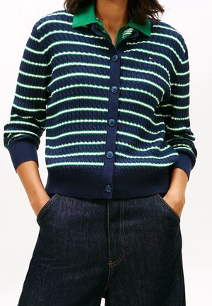Femme portant un cardigan bleu marine avec des rayures horizontales blanches et vertes sur une chemise verte à col et un jean bleu foncé, les mains dans les poches.