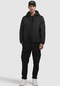 Schwarze Pufferjacke mit hexagonalem Steppmuster, Kapuze und Frontreißverschluss; kombiniert mit schwarzen Cargo-Hosen und weißen Sneakers. Schwarze Kappe getragen.