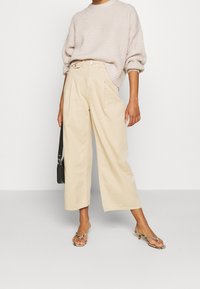 Personne portant un pantalon ample beige, un pull en tricot crème aux manches retroussées, tenant un petit sac à main noir et portant des sandales en cuir de serpent.