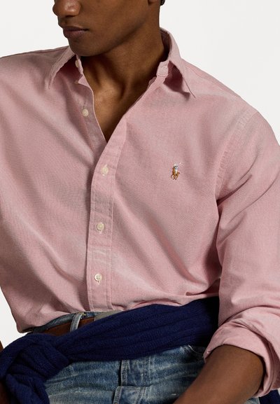 Ljusrosa skjorta med knappar och en subtil textur, prydd med en liten broderad logotyp. Kombinerad med blå jeans och en mörkblå stickad tröja.