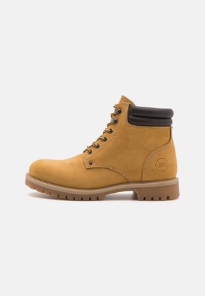 Botas de tobillo de ante color beige con un collar acolchado negro, ojales de metal y cordones, que cuentan con una suela de goma resistente y una marca grabada en el lado.