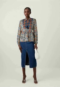 Blusa com padrão floral, de gola redonda e detalhe de laço, combinada com uma saia lápis de ganga com fenda frontal. A modelo segura uma bolsa branca.