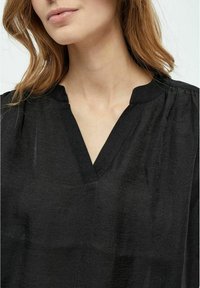 Juoda palaidinė su V-šlo viena, trumpomis rankovėmis ir susiraukšlėtomis detalėmis palei pečius. Audinys turi šiek tiek tekstūruotą išvaizdą.