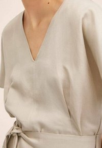 Vêtement beige clair à col en V avec manches courtes, fabriqué en tissu texturé. Il présente une ceinture à nouer à la taille, créant une silhouette décontractée.