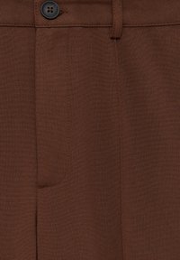 Pantalon marron sur mesure avec une texture de tissu lisse, doté d'une poche avant et d'un bouton noir à la taille.