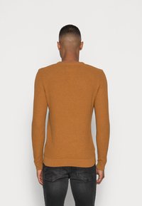 Man som vänder bort med ryggen iklädd en brun texturerad tröja och mörkgrå jeans framför en enkel ljus bakgrund.