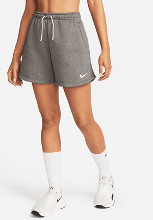 Kobieta w szarych szortach Nike, białych skarpetach sportowych i białych trampkach Nike, stojąca z rękami swobodnie opuszczonymi wzdłuż ciała.