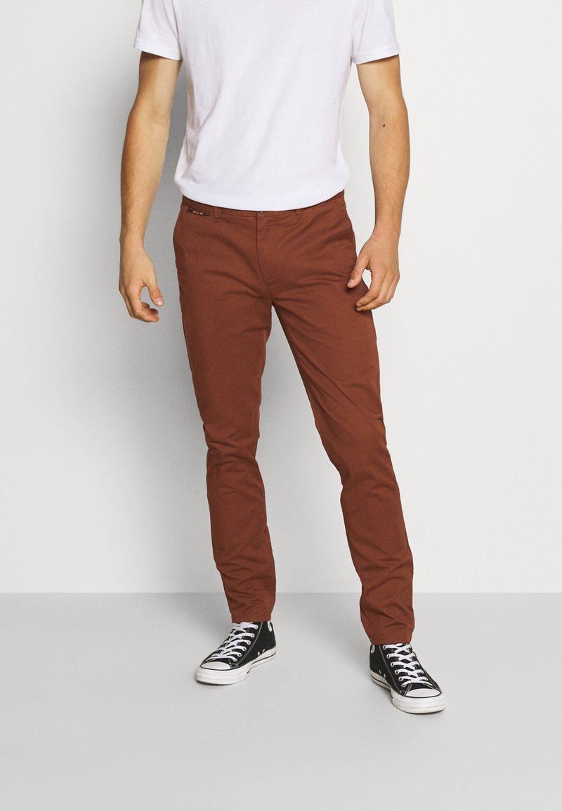 Homme portant un pantalon slim de couleur rouille, une chemise blanche à manches courtes et des baskets montantes noires, se tenant devant un fond uni.