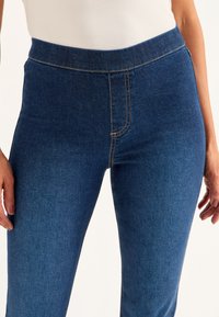 Sinisiä farkkutyylisiä legginssejä, joissa on sileä pinta, korkea vyötärö ja kontrastiväriset oranssit ompeleet etusaumassa. Ei näkyviä taskuja.