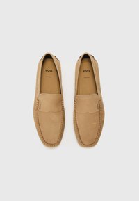 Mocasines de ante en tono marrón claro con textura suave, detalles de costura y punta redondeada. Presenta el logo de la marca impreso en el interior. Diseño de suela plana.