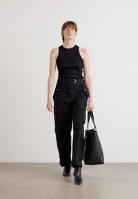 Persona che indossa un top senza maniche nero, pantaloni neri a vita alta, stivaletti neri alla caviglia, mentre tiene una grande borsa tote nera, in piedi contro un muro semplice.