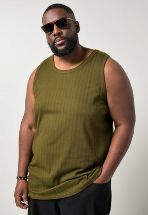 Mann mit Bart und Sonnenbrille, trägt olivgrünes ärmelloses, geripptes Shirt und schwarze Hose, eine Hand in der Tasche, neutrale Hintergrund.