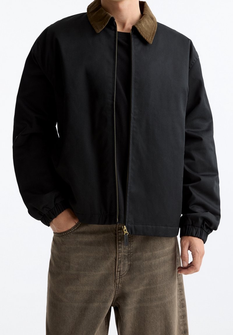 Homme portant une veste zippée noire avec un col marron et des poignets élastiques, associée à un pantalon marron, mains partiellement dans les poches.