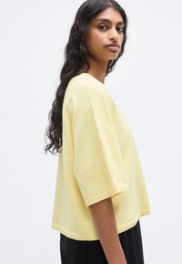 Pull à manches courtes en tissu doux de couleur jaune pâle. Coupe carrée avec col rond et finitions côtelées. Surface légèrement texturée.