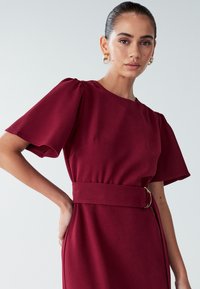 Burgundijas kleita ar īsām, pūkainām piedurknēm, apaļu dekoltē un jostasvietu ar jostu, kas rotāta ar zelta krāsas detaļu. Gluda auduma tekstūra.