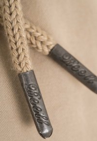 Primer plano de los cordones trenzados beige de una sudadera con capucha, con terminales metálicos grabados con la palabra "Diesel" sobre fondo de tela.