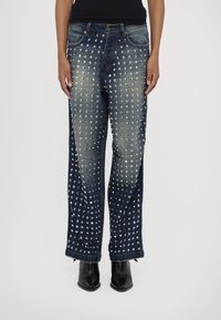 Personne portant un jean en denim bleu foncé orné de clous carrés argentés disposés en motifs verticaux, associé à des bottes noires à bouts pointus.