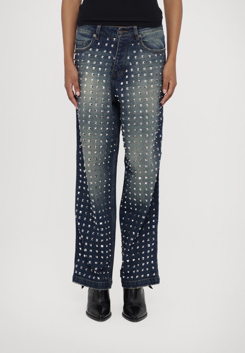 Personne portant un jean en denim bleu foncé orné de clous carrés argentés disposés en motifs verticaux, associé à des bottes noires à bouts pointus.