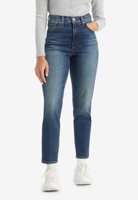 Femme portant un jean bleu taille haute avec un effet délavé à l'avant, un haut à manches longues gris clair, des chaussettes blanches et des baskets blanches, debout avec une main dans la poche.