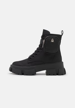 Botte noire à semelle chunky avec lacets, petite pochette zippée latérale avec logo métallique, tirette au talon et semelle robuste à crampons.