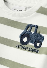 Camisa de algodón blanca con rayas horizontales en verde y gris. Presenta un tractor azul texturizado bordado con la frase "VAMOS A EMPEZAR" debajo.