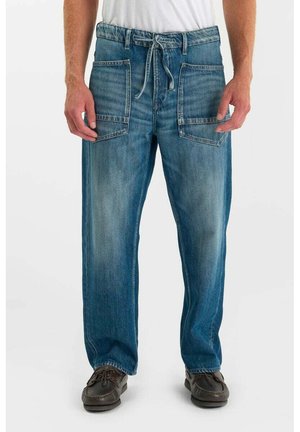 Man draagt blauwe spijkerbroek met grote voorzakken en een tailleband met veter, gecombineerd met donkere loafers.