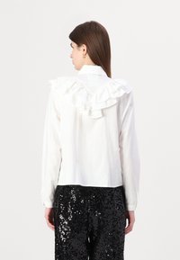 Camisa blanca de manga larga con un cuello con volantes en la parte posterior, combinada con pantalones negros con lentejuelas. Tela suave, corte clásico, sin patrones visibles.