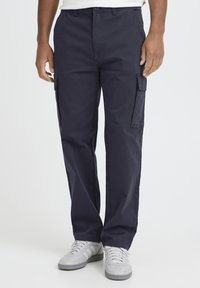 Temno modre cargo hlače iz trpežnega blaga, z bočnimi žepi, ravnimi hlačnicami in zapiranjem na gumb. Nošene z sivimi supergami.