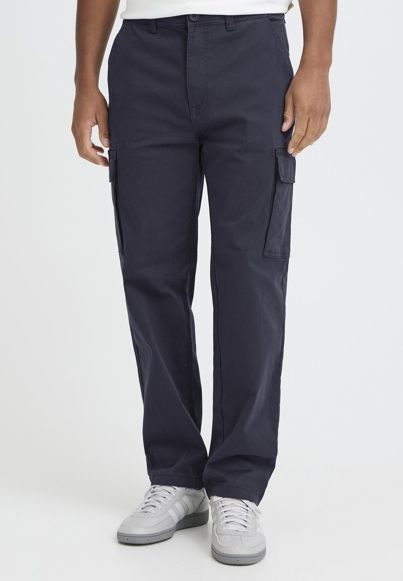 Temno modre cargo hlače iz trpežnega blaga, z bočnimi žepi, ravnimi hlačnicami in zapiranjem na gumb. Nošene z sivimi supergami.