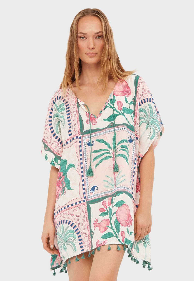 Femme portant une robe tunique ample à manches courtes avec des imprimés botaniques et des glands verts le long de l'ourlet sur un fond uni.