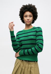 Πράσινο πουλόβερ με πλεκτό σχέδιο καλωδίου, με ναυτικό οριζόντιο ρίγες, στρογγυλή λαιμόκοψη και ριμπ τελειώματα στα μανίκια. Συνδυάζεται με πλισέ μπεζ φούστα.
