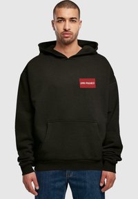 Melns kapucis, izgatavots no mīksta auduma, ar priekšējo kabatu, lielu kapuci un sarkanu taisnstūra formas logo uzšuvumu ar uzrakstu "DIMA MAGHREB".