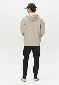 Beige hoodie med en kængurulomme og teksturerede manchetter, båret med mørke jeans og sort-hvide sneakers. Bagview, ensfarvet baggrund.