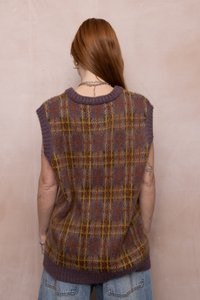 Person med langt rødt hår iført en ærmeløs lilla og brun rutet strikvest samt blå jeans, vendt mod en beige væg.