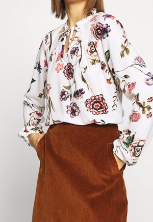 Bloemige blouse in het wit met verschillende kleuren, met een keyhole halslijn en een gerimpelde kraag, gecombineerd met een bruine ribstof rok met zakken.