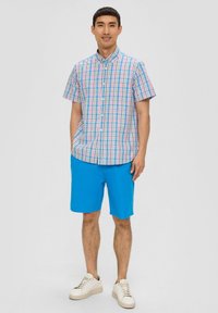 Kurzarmhemd mit Knöpfen in mehrfarbigem Karo (rosa, blau, grün) kombiniert mit einfarbigen blauen Shorts und cremefarbenen Sneakern.