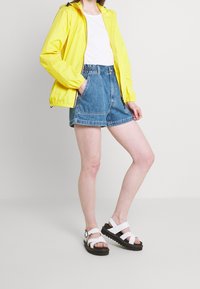 Keltainen vedenpitävä takki, valkoinen t-paita, farkkushortsit ja valkoiset sandaalit mustilla pohjilla. Vapaasti istuva, rento tyyli ja kevyet materiaalit.