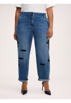 Kvinde iført blå slidte jeans med op-rullede manchetter, sorte pletter og sorte højhælede sko, stående mod en lys baggrund.