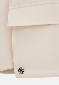 Béžová látka s texturovaným povrchem, se složeným rohem a černým logo knoflíkem na dolním okraji.