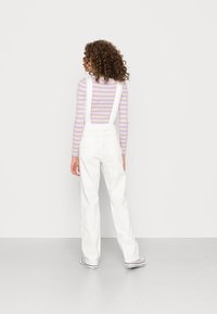 Macacão branco com alças rendadas, combinado com uma camisa de manga longa listrada em tons pastel. Cabelo encaracolado visível por trás.