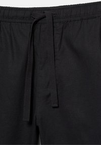 Schwarze Stoffhose mit elastischem Bund und mittiger Kordel, die zu einem lockeren Knoten gebunden ist, mit detaillierter Nahtführung und Textur.