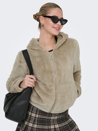 Beige faux fur zip-up hoodie with a relaxed fit, ribbed cuffs, and hem. Worn with a plaid skirt and holding a black bag.