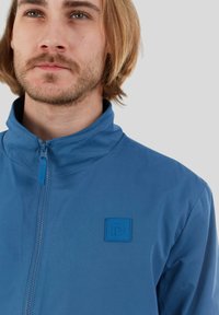 Blaue Zip-Up-Jacke mit hohem Kragen, strukturiertem Stoff und rechteckigem Logopatch auf der linken Seite. Leichtes Design, glatte Oberfläche.