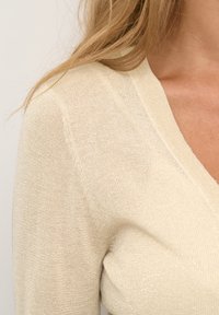 Beige, ribbed knit-sweateri, jossa on V-pääntie ja hienovarainen kimallus. Pehmeä rakenne, istuva malli ja kapeat hihat näkyvät olkapäältä.