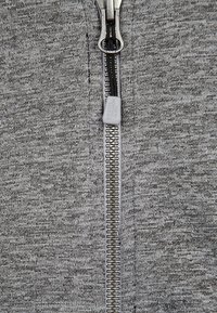 Šedá mikina na zip vyrobená z texturované tkaniny, s kovovým zipem a plastovým poutkem. Jednoduchý design bez vzorů a akcentů.