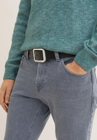 Black leather belt with a square silver buckle, worn over blue-gray denim jeans and a textured teal knit sweater. Close-up view.